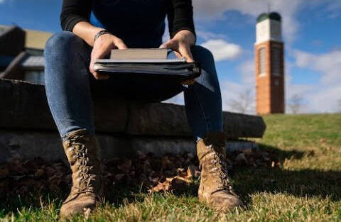 Cut off view of someone sitting, wearing combat boots and holding a binder, with the GVSU clocktower in the background